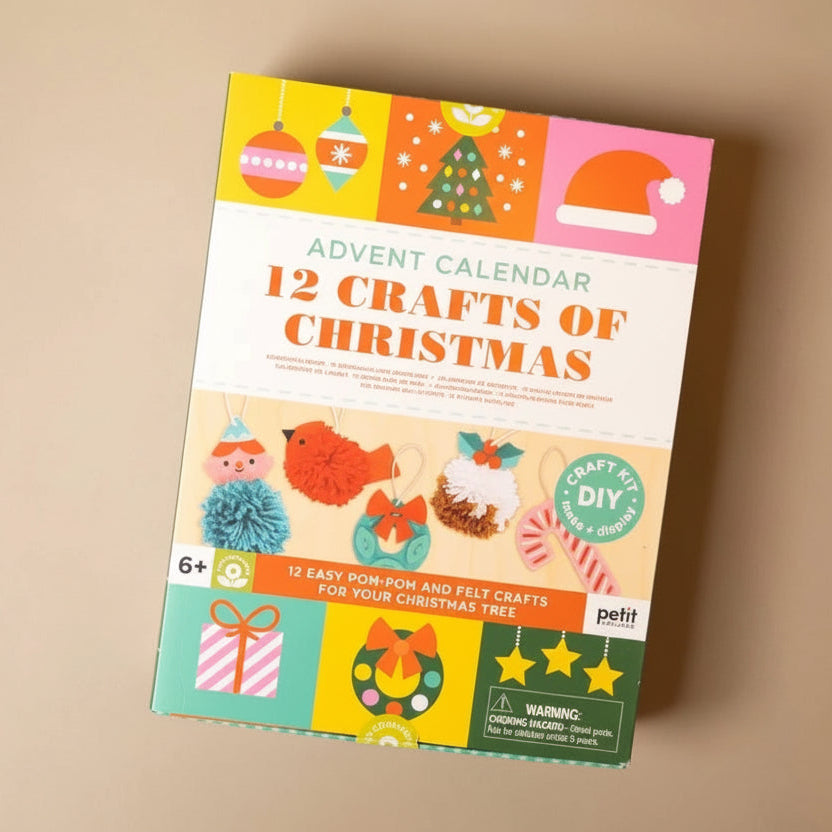 A boxed craft kit titled "Advent Calendar 12 Crafts of Christmas" photographed on a warm beige background. The package is angled slightly and includes a hanging paper tag with a silhouette figure.