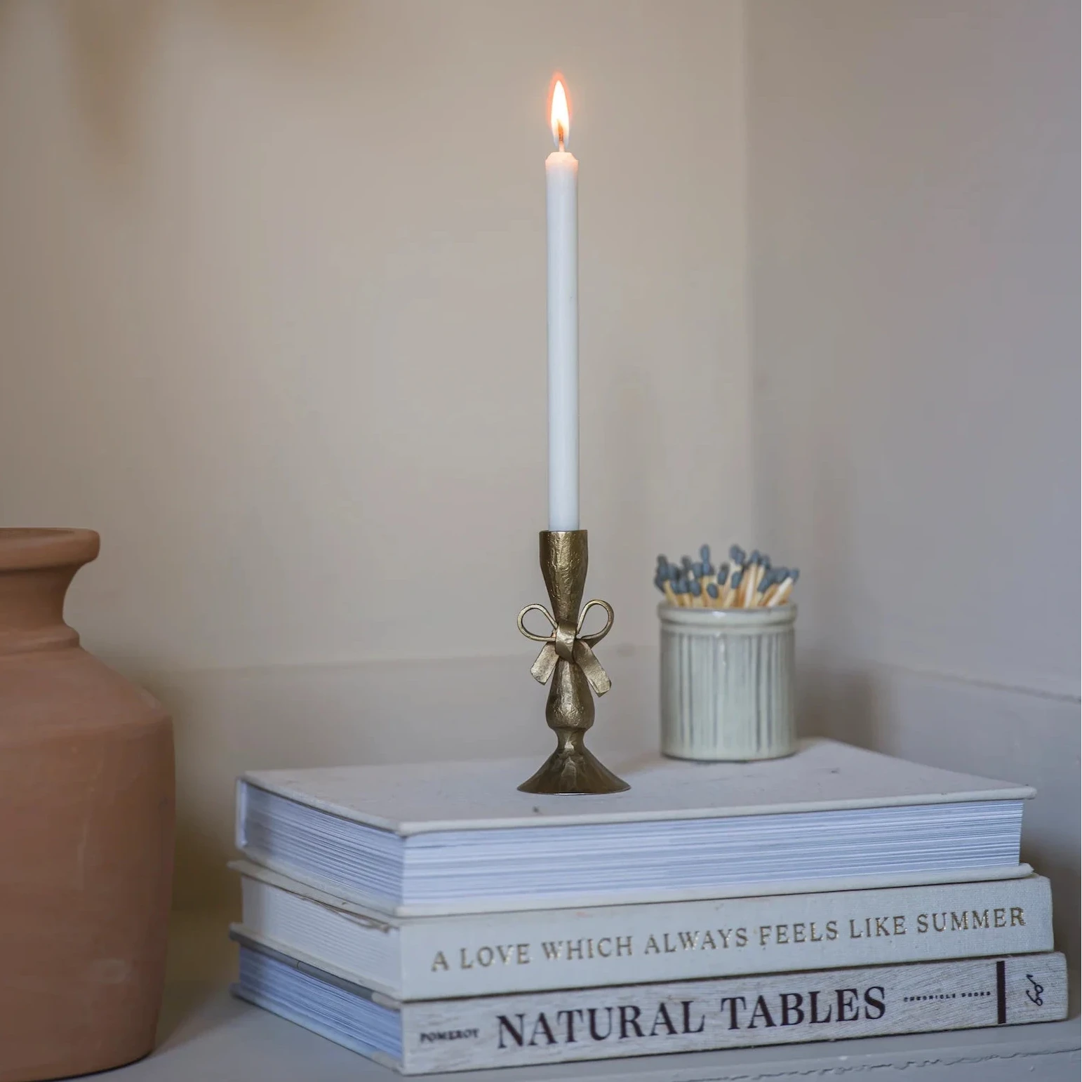 Candle holder in brushed antique brass with a bow detail, sat on a stacked pile of books with a burning dinner candle inside, reminiscent of a Victorian scene.