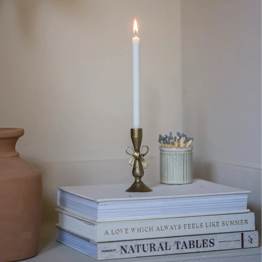 Candle holder in brushed antique brass with a bow detail, sat on a stacked pile of books with a burning dinner candle inside, reminiscent of a Victorian scene.