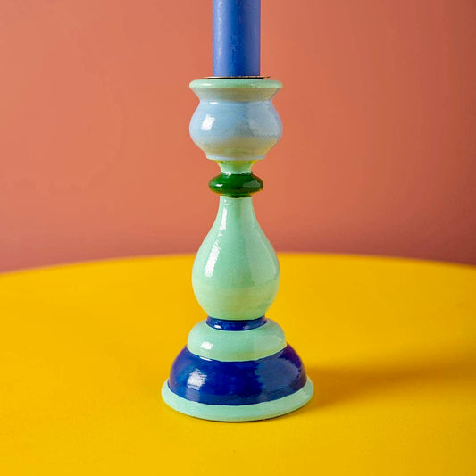 Ian Snow iconic shaped candle holder in light blue with dark green and cobalt contrasting rings. Featured holding a bright blue candle on a contrasting yellow surface and warm pink background. Part of SW Coast's Merry & Bright Christmas Collection.