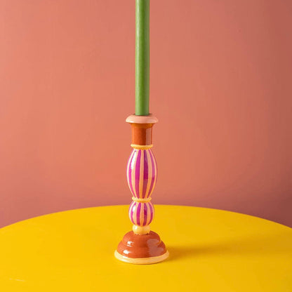 Ian Snow iconic shaped candle holder in brick red with pink and yellow trim and a striking fairground pink candy striped stem.  Featured holding a bright green candle on a contrasting yellow surface and warm pink background. Part of SW Coast's Merry & Bright Christmas Collection.