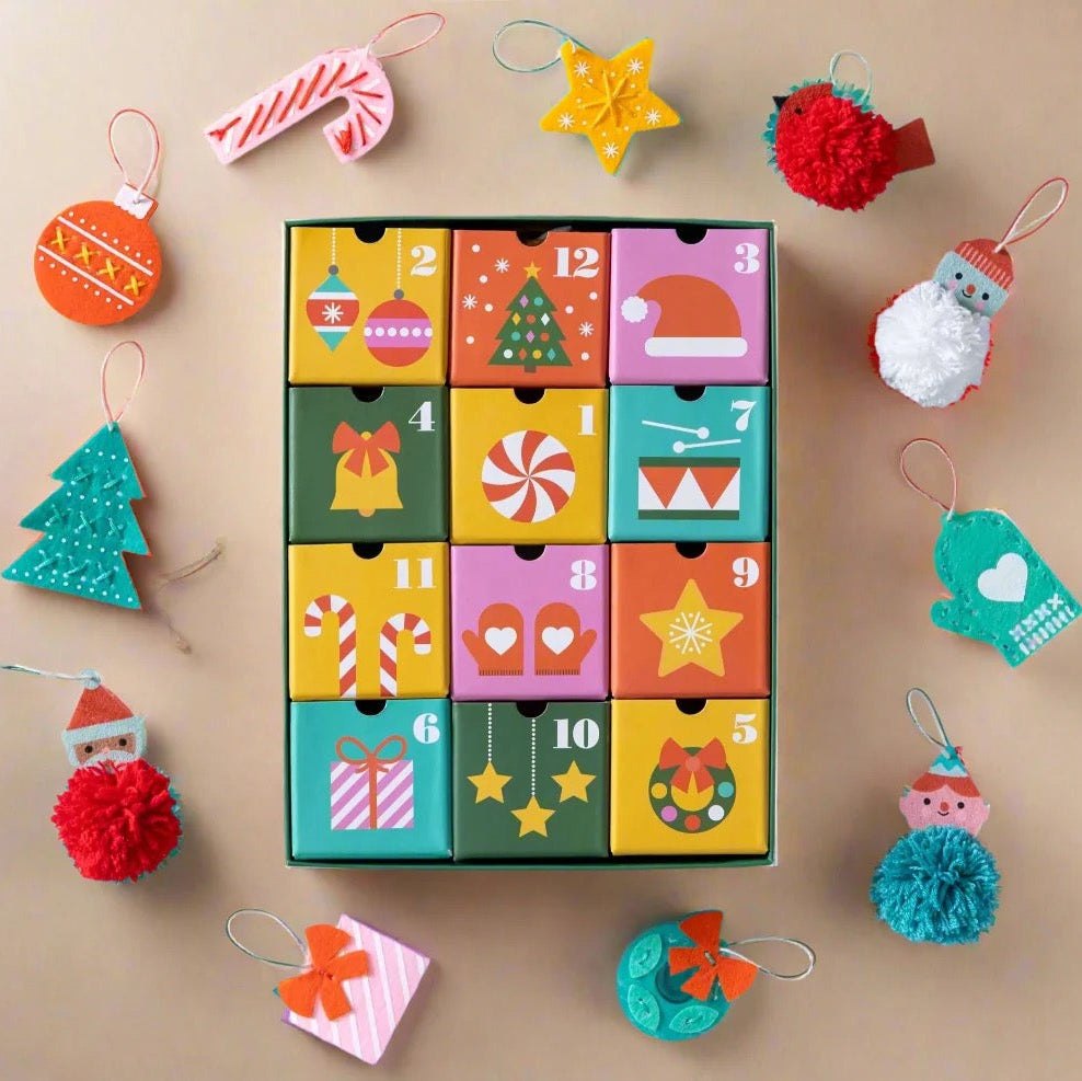 A colorful Christmas advent calendar with 12 numbered drawers arranged in a grid, surrounded by festive hanging ornaments on a beige background.