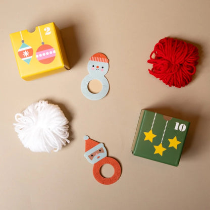 A Christmas-themed sensory craft kit arranged on a beige background, featuring two wooden snowman rings, a yellow box with hanging ornament illustrations and the number 2, a green box with hanging star illustrations and the number 10, a red ball of yarn, and a white pom-pom.