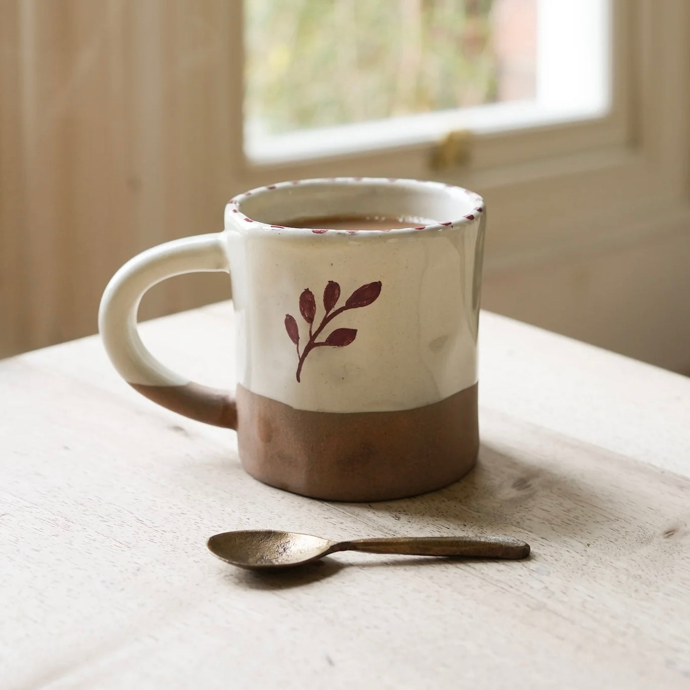 Hand painted natural stoneware ceramic mug with a botanic leaf design on a wooden surface next to an antique brass spoon. Rust Red Hand Painted Botanic Ceramic Mug | The Perfect Gift for Coffee Lovers