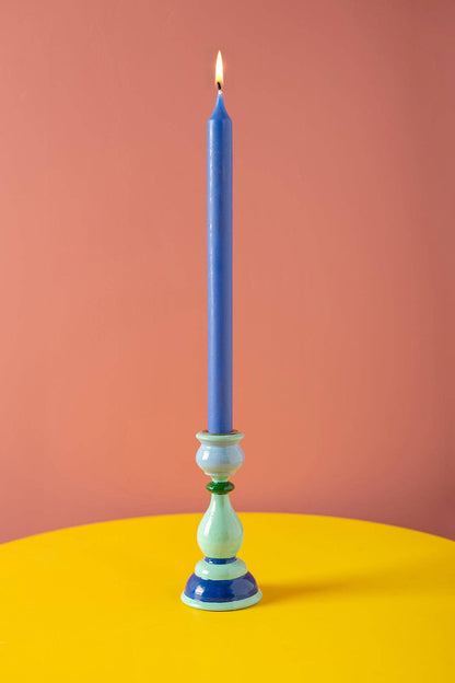 Blue candle displayed lit in a pale blue decorative candlestick with navy blue and dark green bands against a pink background.